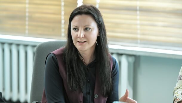 Moteris sėdi prie stalo ir gestikuliuoja dalykinio susitikimo metu