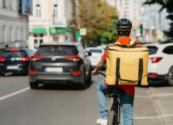 Dviratininkas su dideliu pristatymo krepšiu važiuoja miesto gatve tarp automobilių