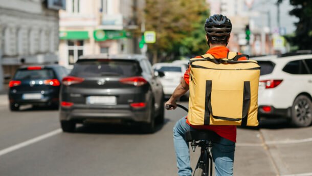 Dviratininkas su dideliu pristatymo krepšiu važiuoja miesto gatve tarp automobilių