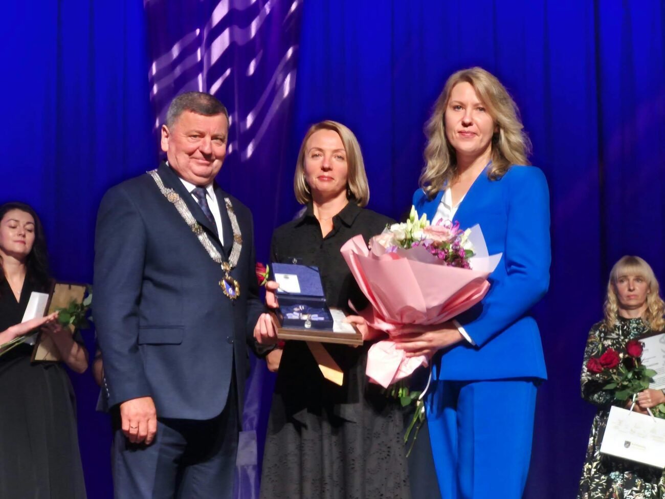 Apdovanojimo ceremonijoje trys žmonės scenoje laiko gėles ir įvertinimą