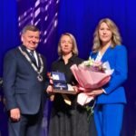 Apdovanojimo ceremonijoje trys žmonės scenoje laiko gėles ir įvertinimą