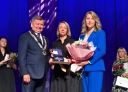 Apdovanojimo ceremonijoje trys žmonės scenoje laiko gėles ir įvertinimą