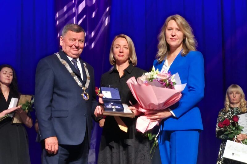 Apdovanojimo ceremonijoje trys žmonės scenoje laiko gėles ir įvertinimą