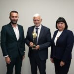 Trys oficialiai apsirengę žmonės pozuoja, o viduryje vyras laiko apdovanojimą