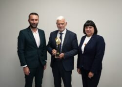 Trys oficialiai apsirengę žmonės pozuoja, o viduryje vyras laiko apdovanojimą