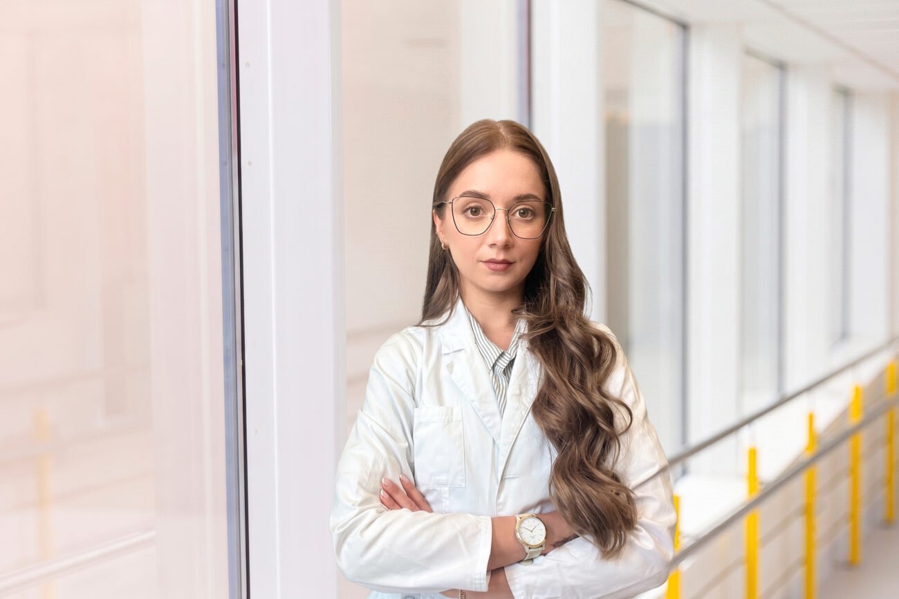 Baltais chalatais vilkinti medicinos specialistė stovi ligoninės koridoriuje sukryžiuotomis rankomis