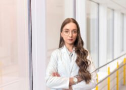 Baltais chalatais vilkinti medicinos specialistė stovi ligoninės koridoriuje sukryžiuotomis rankomis