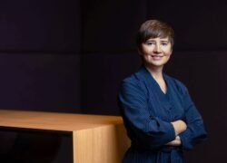 Moteris mėlynais marškiniais sukryžiuotomis rankomis stovi prie medinio stalo