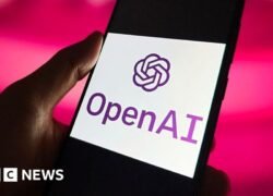 Rankoje laikomas išmanusis telefonas su ekrane rodomu OpenAI logotipu ir pavadinimu