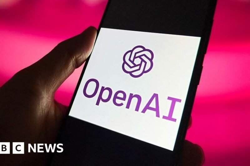 Rankoje laikomas išmanusis telefonas su ekrane rodomu OpenAI logotipu ir pavadinimu