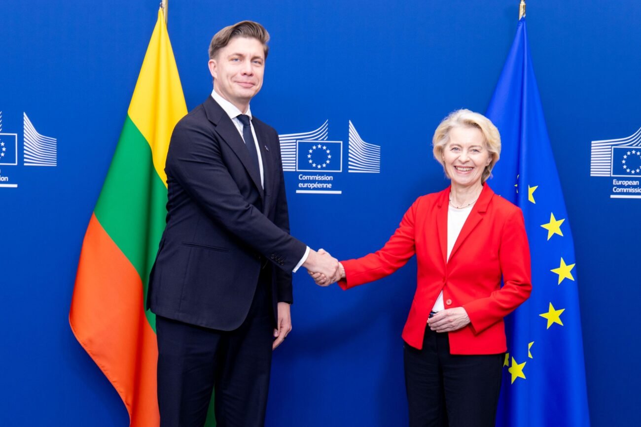 Du žmonės spaudžia rankas Europos Komisijos ženklu pažymėtame fone