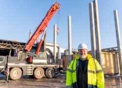 Statybų aikštelėje darbuotojas stovi šalia krano ir betoninių kolonų