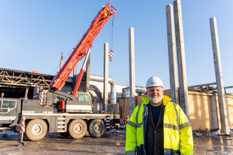 Statybų aikštelėje darbuotojas stovi šalia krano ir betoninių kolonų
