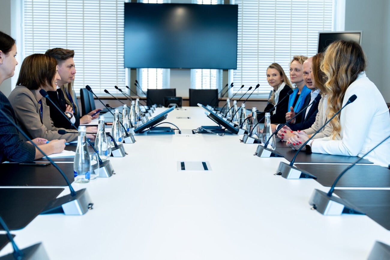 Dalyviai susėdę prie konferencijų stalo su mikrofonais ir ekranu