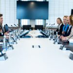 Dalyviai susėdę prie konferencijų stalo su mikrofonais ir ekranu