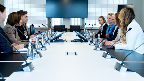Dalyviai susėdę prie konferencijų stalo su mikrofonais ir ekranu