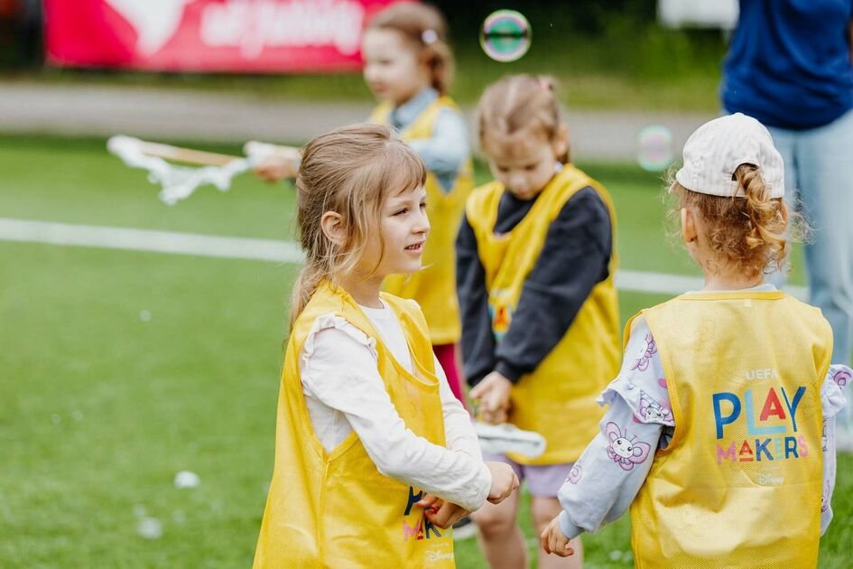 Vaikai geltonomis liemenėmis žaidžia futbolo aikštėje
