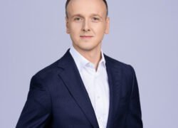 Vyras su tamsiai mėlynu kostiumu ir baltais marškiniais šviesiame fone