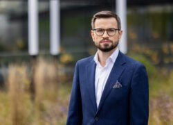 Barzdotas vyras su akiniais stovi lauke priešais modernų pastatą