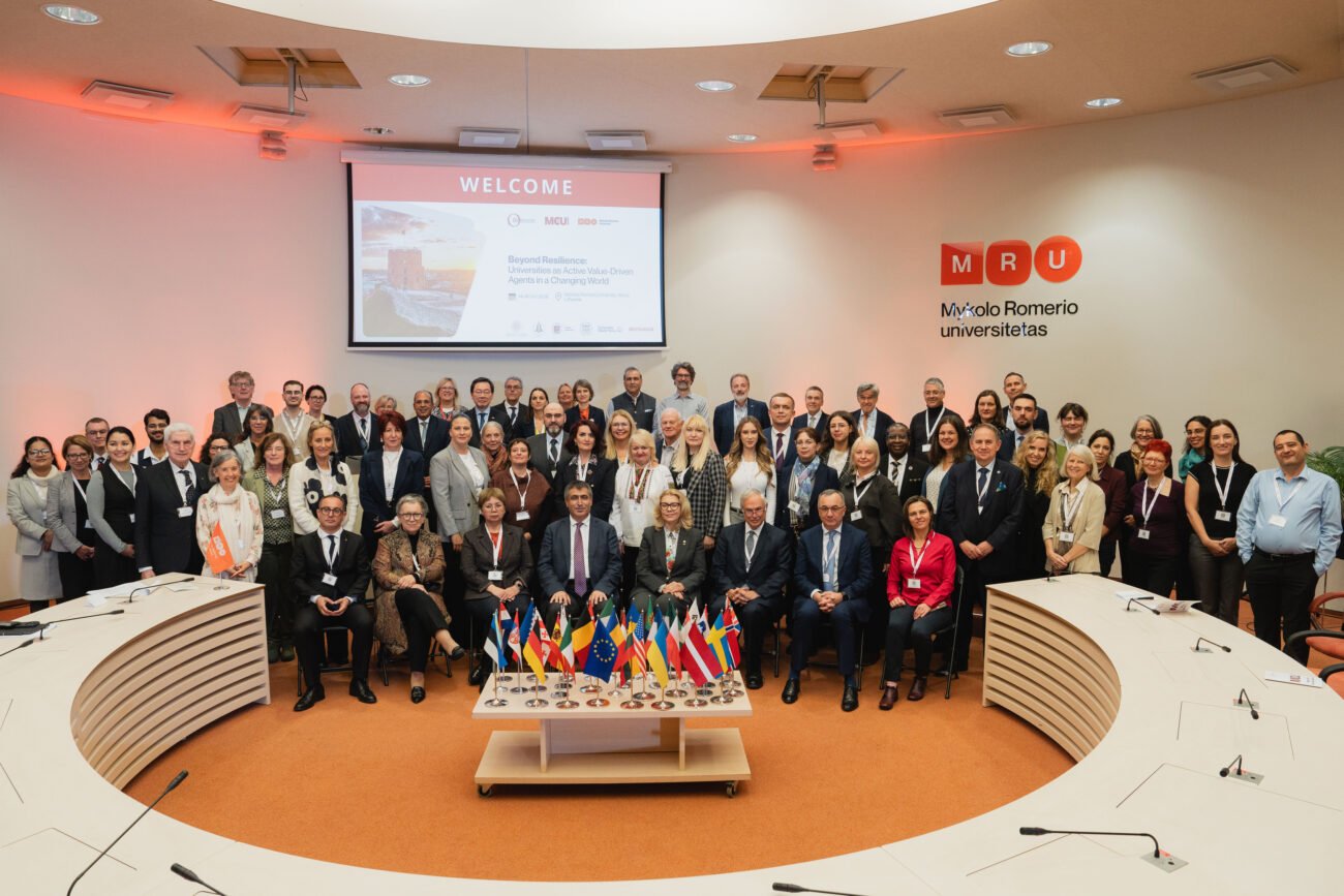 Konferencijos dalyvių grupė posėdžių salėje prie stalo ir tarptautinių vėliavėlių