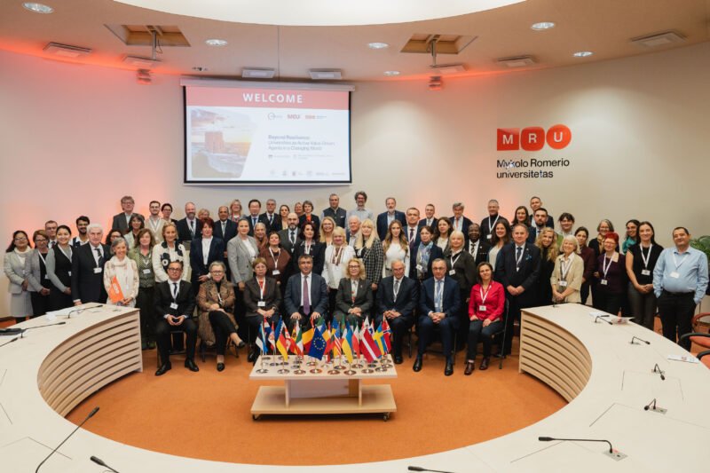 Konferencijos dalyvių grupė posėdžių salėje prie stalo ir tarptautinių vėliavėlių