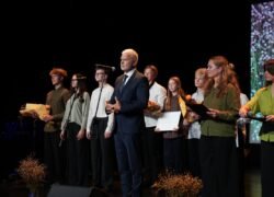 Scenoje stovi grupė žmonių, o priekyje kalba kostiumuotas vyras