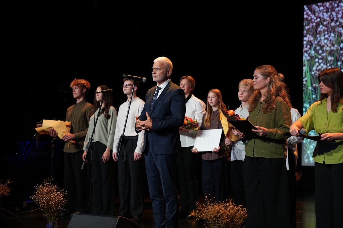 Scenoje stovi grupė žmonių, o priekyje kalba kostiumuotas vyras
