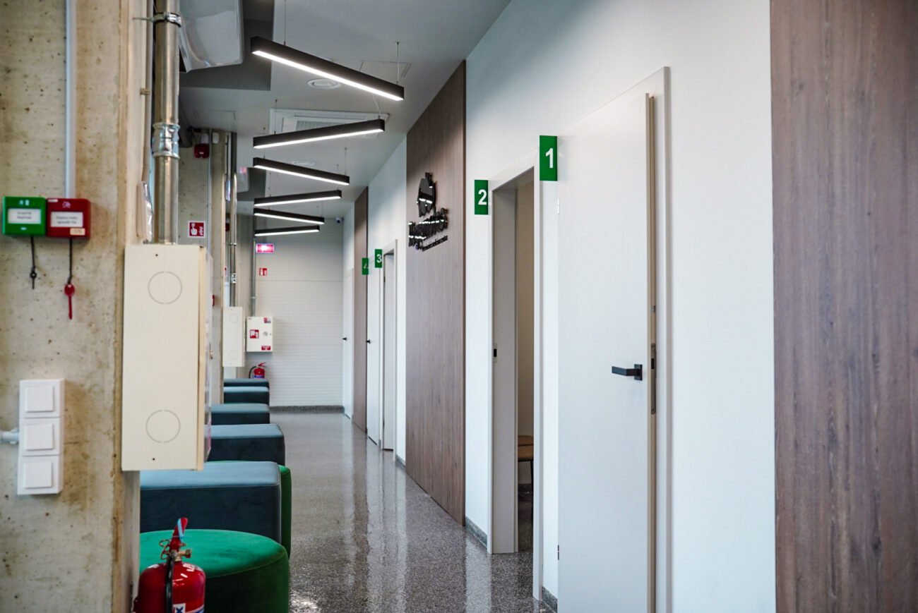 Šviesus klinikos koridorius su numeruotomis durimis, laukimo vietomis ir gesintuvu
