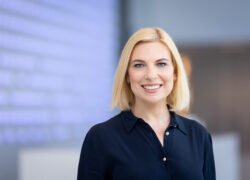 Besišypsanti blondinė moteris tamsiais marškiniais biuro aplinkoje