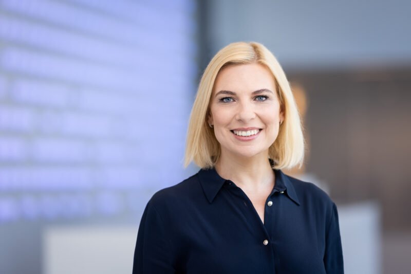 Besišypsanti blondinė moteris tamsiais marškiniais biuro aplinkoje