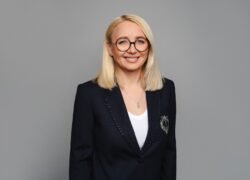 Besijuokianti šviesiaplaukė moteris su akiniais ir tamsiu švarku pilkame fone