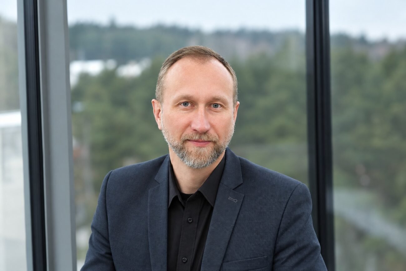 Barzdotas vyras su tamsiu švarku stovi priešais langą
