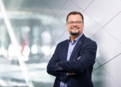 Akiniais pasipuošęs vyras sukryžiuotomis rankomis stovi moderniame biure
