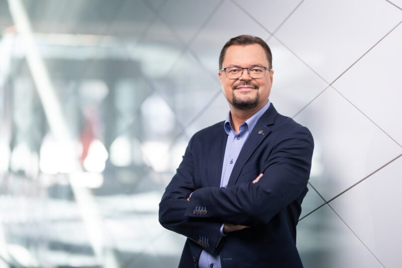 Akiniais pasipuošęs vyras sukryžiuotomis rankomis stovi moderniame biure