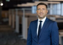 Mėlynu kostiumu pasipuošęs vyras stovi moderniame biure