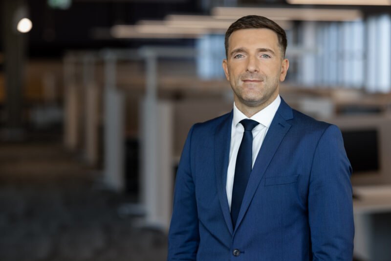Mėlynu kostiumu pasipuošęs vyras stovi moderniame biure