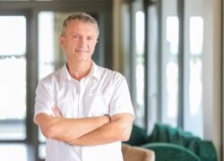 Baltus marškinius vilkintis vyras sukryžiuotomis rankomis šviesiame interjere