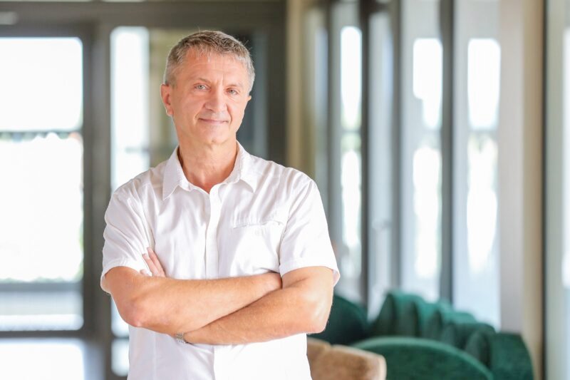 Baltus marškinius vilkintis vyras sukryžiuotomis rankomis šviesiame interjere