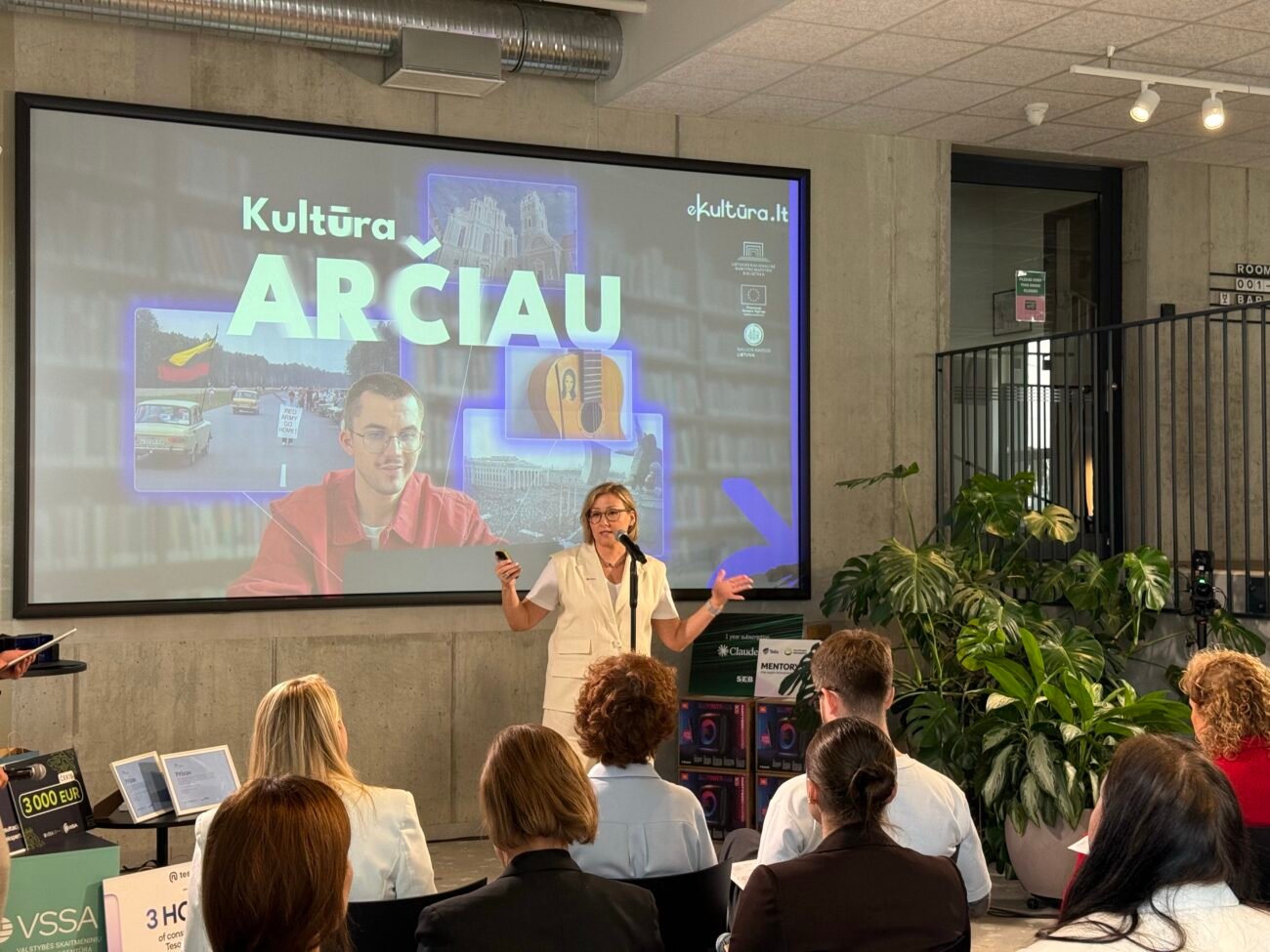 Moteris pristato kultūros platformą auditorijai, ekrane matomas užrašas „Kultūra ARČIAU“