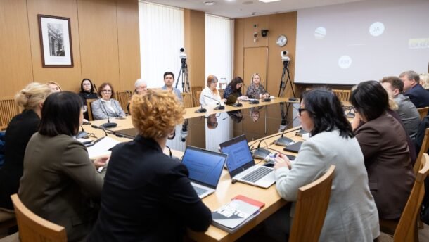 Dalyviai susėdę prie stalo diskutuoja konferencijų salėje