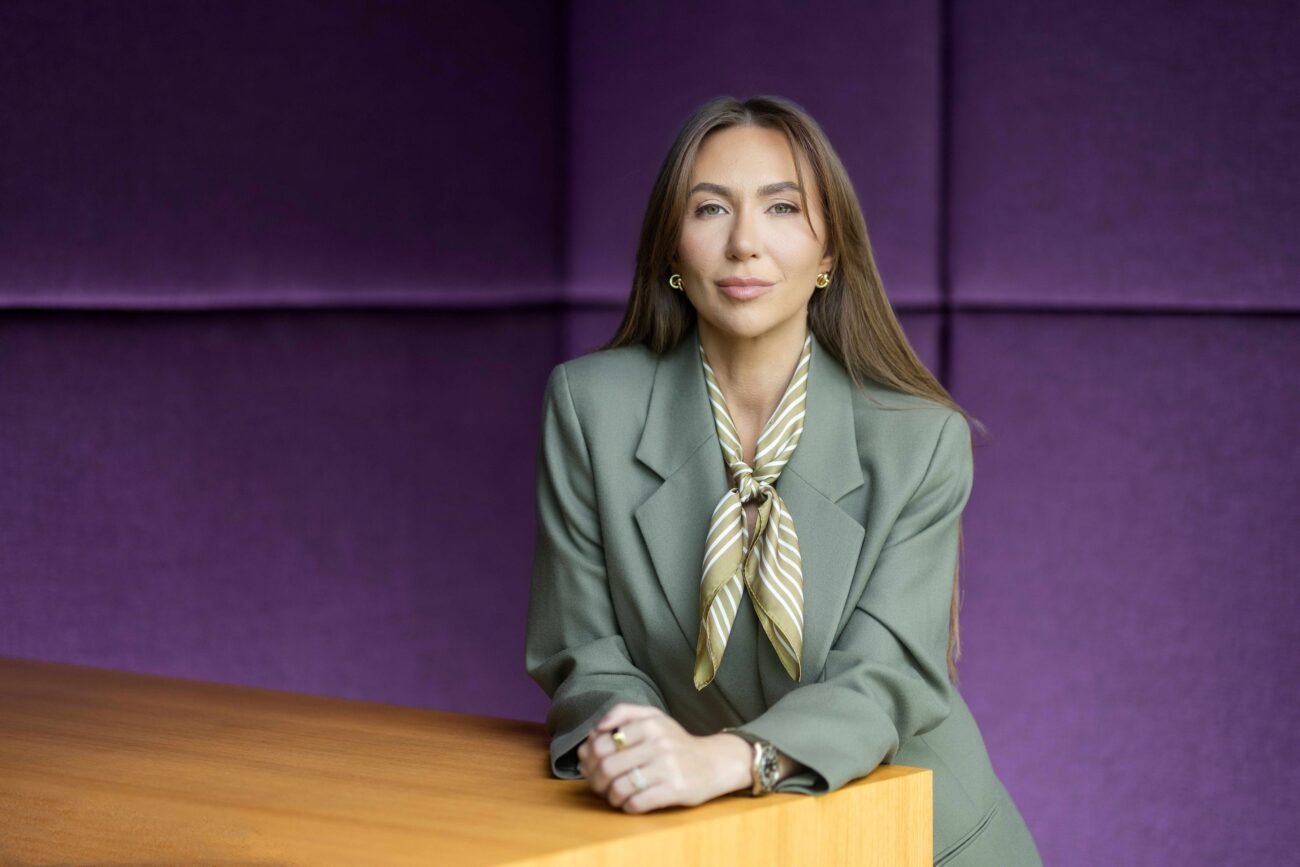 Ieva Linkutė - Urbonė sėdi prie medinio stalo, vilkėdama pilką kostiumą ir šaliką.