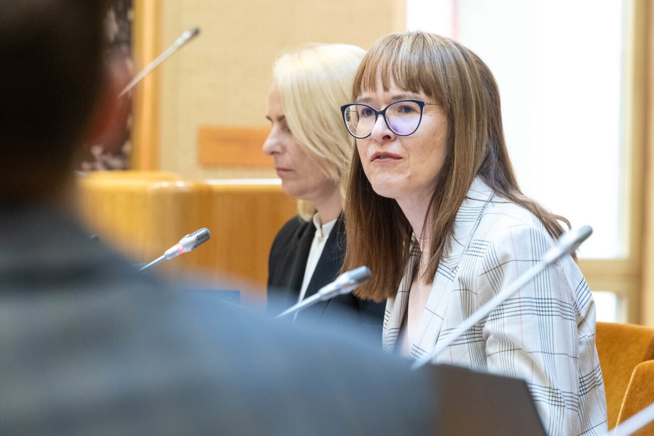 Dvi moterys posėdžių salėje sėdi prie mikrofonų