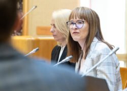 Dvi moterys posėdžių salėje sėdi prie mikrofonų