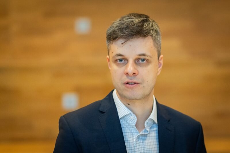 Jaunas vyras su tamsiu švarku pozuoja medinės sienos fone