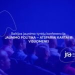 Mėlyna šviesa apšviesti konferencijos dalyviai sėdi auditorijoje