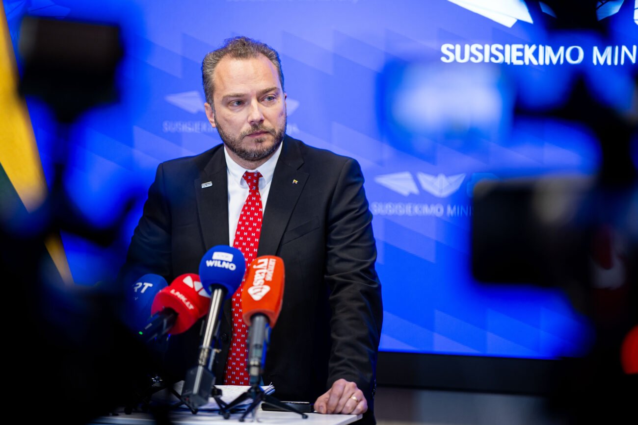 Kostiumuotas vyras prie mikrofonų Susisiekimo ministerijos fone