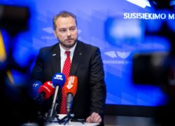 Kostiumuotas vyras prie mikrofonų Susisiekimo ministerijos fone