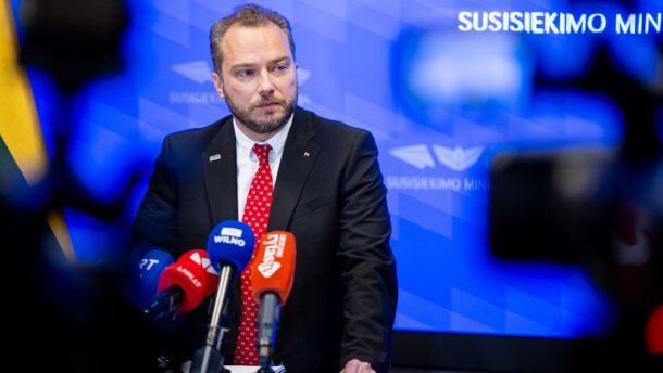 Kostiumuotas vyras prie mikrofonų Susisiekimo ministerijos fone
