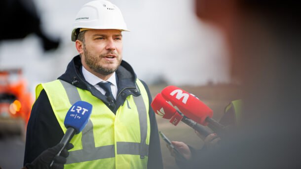 Statybos sektoriaus atstovas su šalmu ir liemene kalba žurnalistams