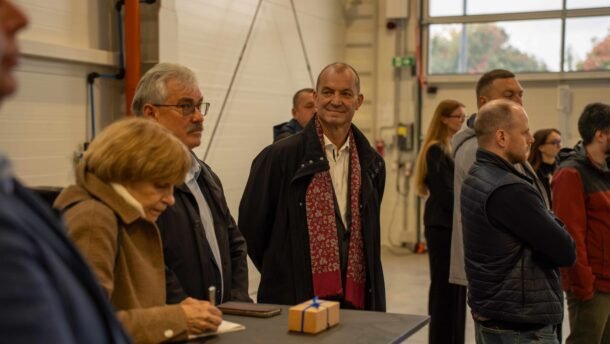 Žmonės susirinkę pramoninėse patalpose, ant stalo matoma maža dovanų dėžutė